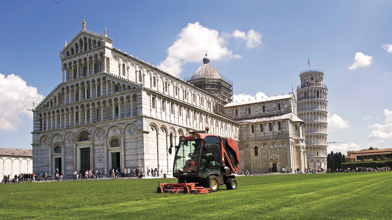 Large, orange lawn mower cutting grass in front of a crowded church